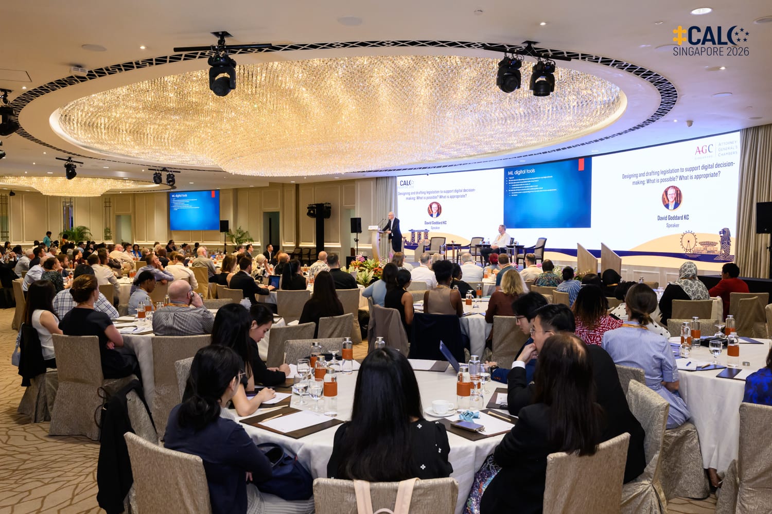 A wide shot of the ballroom at The Fullerton Hotel Singapore during a Conference session, with delegates listening to speaker David Goddard KC at the podium.